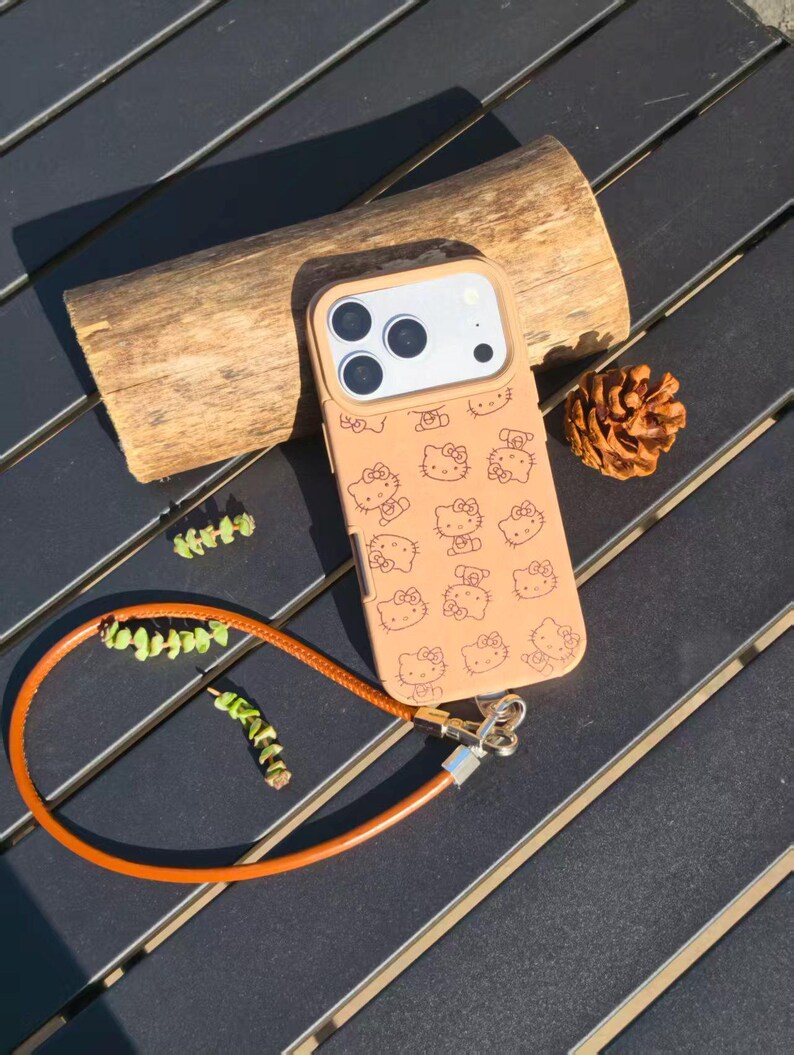 May include: A tan smartphone case featuring a repeating pattern of Hello Kitty character outlines. The case includes a camera cutout and a detachable orange wrist strap with a silver metal clasp, providing a secure way to carry the device.
