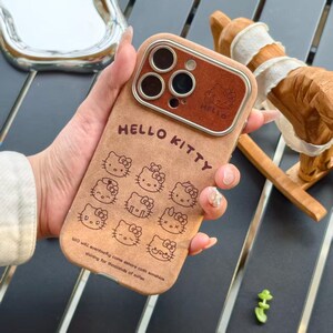 Op de afbeelding: Een bruin telefoonhoesje met Hello Kitty-ontwerpen. De hoes heeft een bruine cameralensbeschermer en de woorden "HELLO KITTY" staan bovenaan. De achterkant van de hoes heeft meerdere Hello Kitty-illustraties en tekst onderaan.