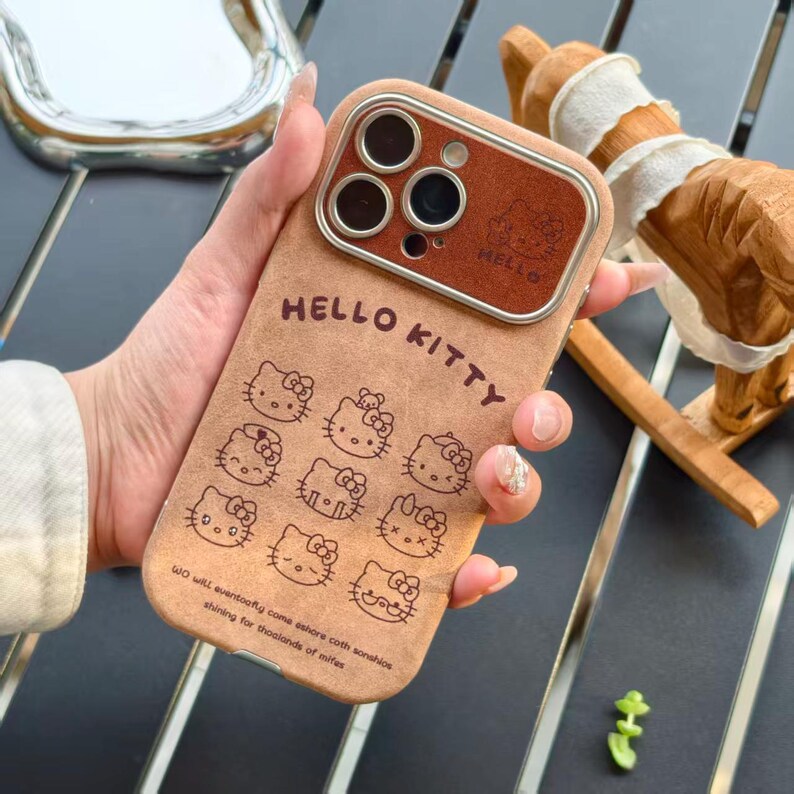Op de afbeelding: Een bruin telefoonhoesje met Hello Kitty-ontwerpen. De hoes heeft een bruine cameralensbeschermer en de woorden "HELLO KITTY" staan bovenaan. De achterkant van de hoes heeft meerdere Hello Kitty-illustraties en tekst onderaan.