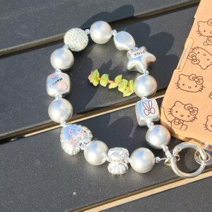 May include: A beaded bracelet featuring large, round, and irregular silver beads. The bracelet includes charms with a strawberry, a star with "PINK" text, and a bunny. A small sprig of green leaves is next to the bracelet.