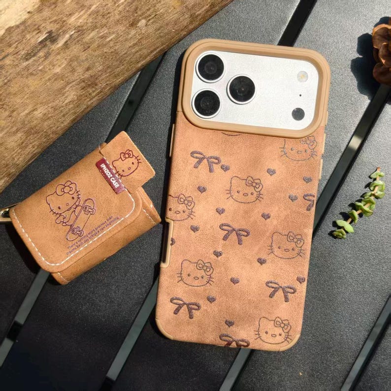 K&ouml;nnte beinhalten: Braune Handyh&uuml;lle und passendes Schl&uuml;sseletui mit einem sich wiederholenden Muster aus Hello Kitty-Gesichtern und Schleifen. Die Handyh&uuml;lle hat einen Kameraausschnitt und einen beigen Rand. Das Schl&uuml;sseletui hat einen Druckknopfverschluss.