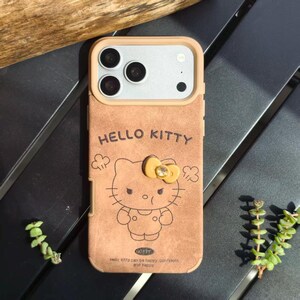 May include: A tan phone case featuring a Hello Kitty design. The case has a tan frame and a Hello Kitty graphic with a yellow bow. The text "HELLO KITTY" is above the graphic, and the text "Hello Kitty can be happy, confident and happy" is below.