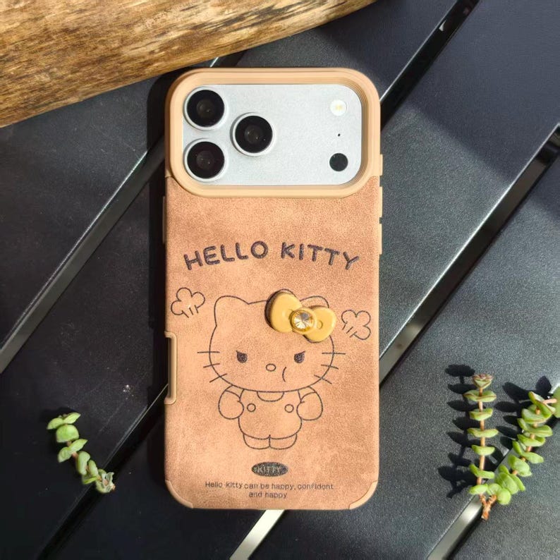 May include: A tan phone case featuring a Hello Kitty design. The case has a tan frame and a Hello Kitty graphic with a yellow bow. The text "HELLO KITTY" is above the graphic, and the text "Hello Kitty can be happy, confident and happy" is below.