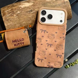 May include: Brown Hello Kitty phone case with matching small pouch. The phone case features a repeating pattern of Hello Kitty faces and bows. The pouch has the words "HELLO KITTY" printed on it. The case and pouch are made of a textured material.