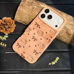 May include: A tan phone case featuring a pattern of Hello Kitty faces, bows, and hearts. The case has a light brown border and a camera cutout. The phone case is on a dark surface with a piece of wood and a pinecone in the background.