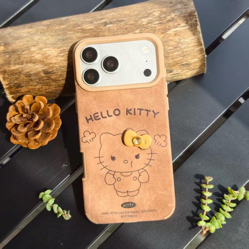 May include: A tan phone case featuring a Hello Kitty design. The case has a light brown frame around the camera and a Hello Kitty graphic with a yellow bow. The text "HELLO KITTY" is above the character. The case also has the text "Hello kitty can be happy, confident and happy".