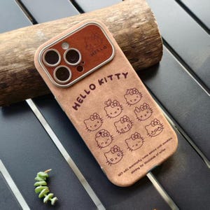 Op de afbeelding: Een bruine telefoonhoes met Hello Kitty-ontwerpen. De hoes heeft een su&egrave;de-achtige textuur en toont de woorden "HELLO KITTY" en meerdere Hello Kitty-gezichten. Het camera gedeelte is donkerder bruin met een zilverkleurige metalen rand.