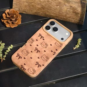 May include: A tan phone case featuring a repeating pattern of Hello Kitty faces, bows, and hearts. The case has a light brown border and a silver camera section. The case is on a dark surface with a pinecone and greenery in the background.