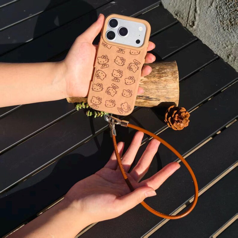 May include: A tan smartphone case featuring a repeating pattern of Hello Kitty character faces in dark brown. The case includes a matching tan wrist strap attached to the bottom with a silver metal clasp, designed for secure carrying of the device.