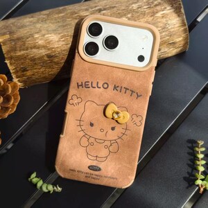May include: Brown phone case featuring a Hello Kitty design. The case has a tan border around the camera and a yellow bow with a gem. The text "HELLO KITTY" is embossed at the top, with additional text at the bottom. The case is designed for a smartphone.