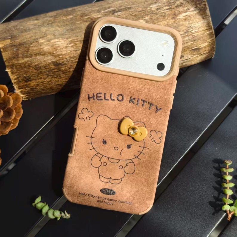 May include: Brown phone case featuring a Hello Kitty design. The case has a tan border around the camera and a yellow bow with a gem. The text "HELLO KITTY" is embossed at the top, with additional text at the bottom. The case is designed for a smartphone.