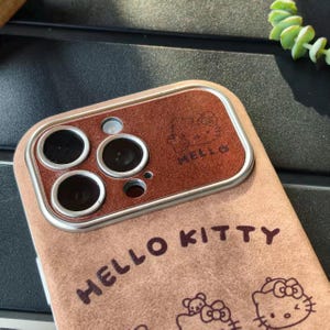 Op de afbeelding: Een bruin telefoonhoesje met de tekst "HELLO KITTY" en een Hello Kitty-afbeelding. De hoes heeft een bruin accent rond de cameralenzen en het woord "HELLO" is in reli&euml;f op het bruine accent.