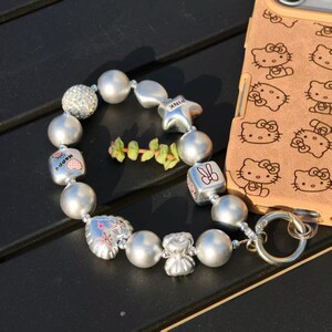 May include: Silver beaded bracelet with various charms, including a star, a cube with a bunny, and a heart with the word "Happy". The bracelet is next to a phone case with a Hello Kitty design.