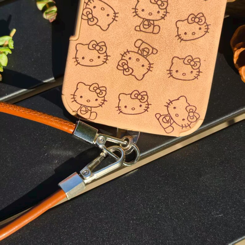 May include: A tan phone case featuring a repeating pattern of Hello Kitty faces in black ink. The case includes a silver metal ring attachment connected to a smooth, brown leather-like strap with silver metal lobster claw clasps.