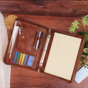 May include: A brown leather-like zippered portfolio organizer featuring a three-ring binder, a grid-lined notepad, multiple card slots with colorful inserts, three pen loops, and a vertical document pocket with a snap-button closure.