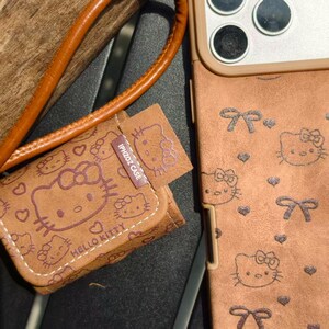 May include: Brown phone case and matching small pouch featuring Hello Kitty designs. The case has a strap and a matching pouch with the text "IPHZOZ CASE" and "HELLO KITTY". The case has a bow and heart pattern.