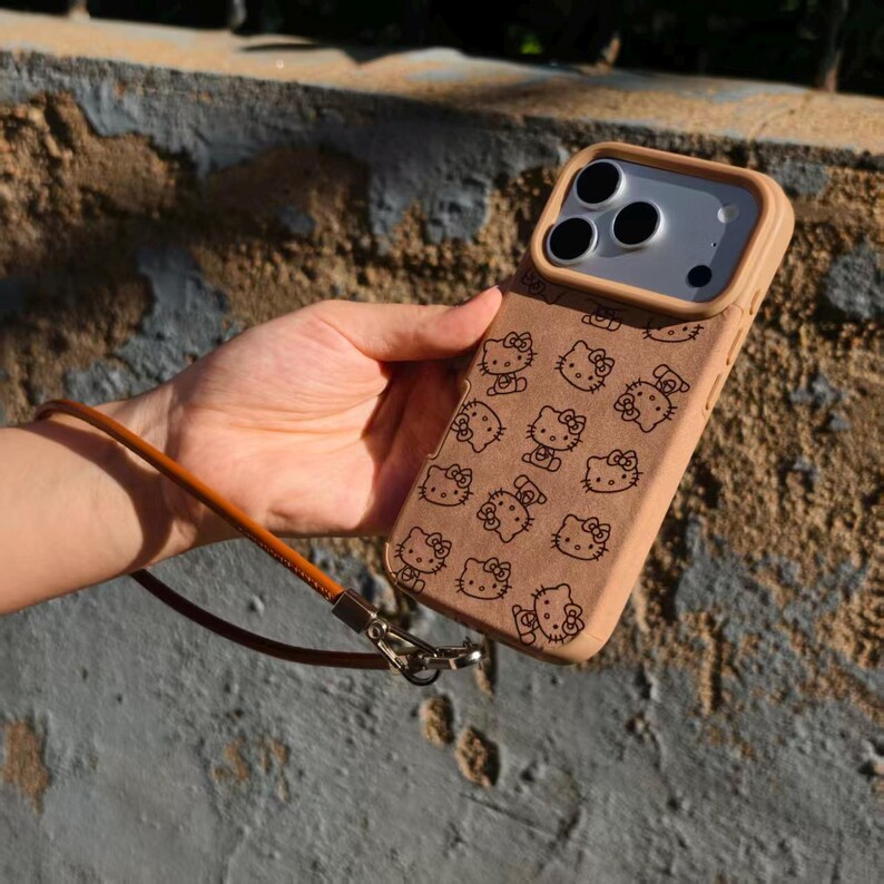 May include: A tan phone case featuring a repeating black line art pattern of Hello Kitty faces. The case includes a raised camera frame and a detachable brown leather wrist strap attached to a silver metal clasp on the side of the device.