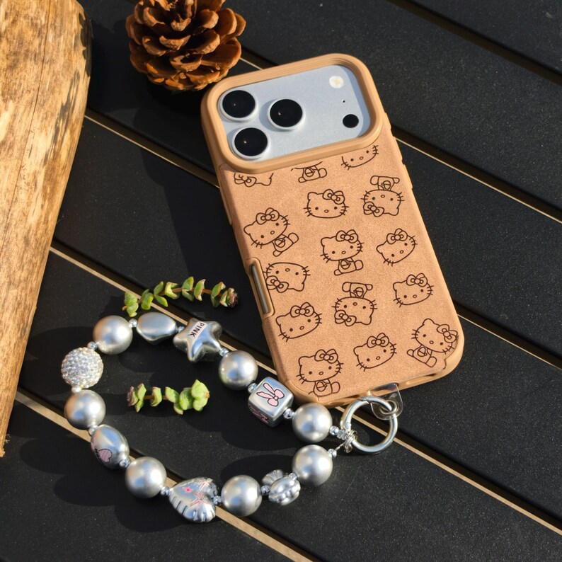 May include: Tan phone case with a repeating pattern of Hello Kitty characters. A silver beaded bracelet with various charms and beads is next to the phone case. The bracelet includes a star charm with the word "PINK" on it.