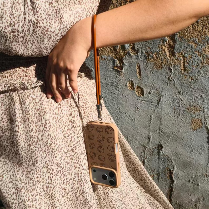 May include: A tan smartphone case featuring a repeating pattern of small cartoon cat faces, attached to a long, thin, brown leather wrist strap. The case has a rectangular cutout for the camera lenses and is suspended from a wrist.