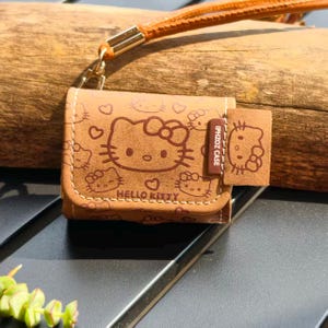 May include: A brown leather case featuring a Hello Kitty design with hearts and the text "HELLO KITTY". The case has a matching tag that reads "IPH2OZ CASE". It includes a brown wrist strap with gold-colored hardware.