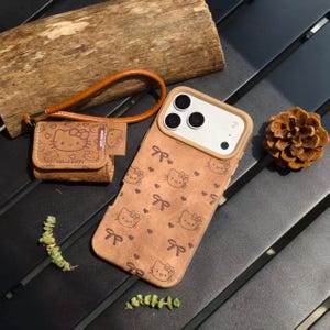 May include: Brown phone case and matching small pouch featuring a repeating pattern of Hello Kitty faces and bows. The phone case has a camera cutout and a brown leather wrist strap. The pouch has a brown leather strap.