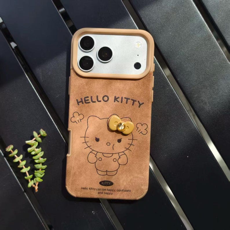 May include: Brown phone case featuring a Hello Kitty design. The case has a tan frame around the camera and a brown, textured back with the words "HELLO KITTY" and a cartoon image of Hello Kitty. The case also has the text "Hello kitty can be happy, confident and happy."