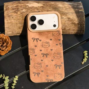May include: A brown phone case featuring a repeating pattern of Hello Kitty faces, bows, and hearts. The case has a tan border around the camera and phone edges. The case is on a dark surface with a piece of wood.