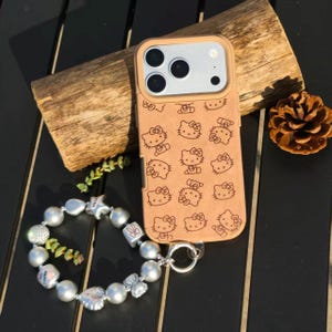 May include: A tan phone case featuring a repeating pattern of Hello Kitty faces. A silver charm bracelet with pearl beads and decorative charms is next to the phone case. The phone case is on a wooden surface.