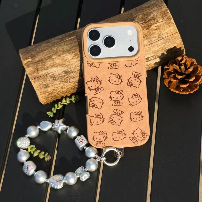 May include: A tan phone case featuring a repeating pattern of Hello Kitty faces. A silver charm bracelet with pearl beads and decorative charms is next to the phone case. The phone case is on a wooden surface.