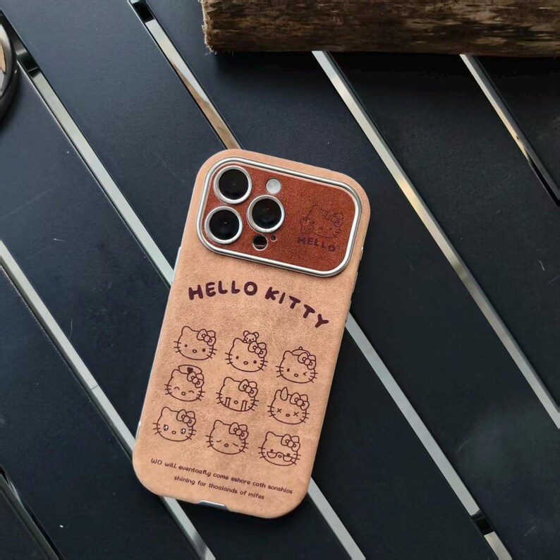 Op de afbeelding: Tan telefoonhoesje met het Hello Kitty-logo en meerdere illustraties van het personage. De hoes heeft een bruin lederlook accent rond de cameralenzen en de tekst "HELLO KITTY" staat op de hoes.