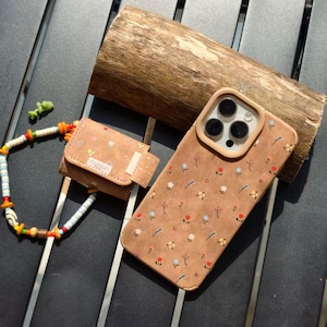 May include: A brown phone case and matching small pouch with a floral pattern. The phone case has a camera cutout and a light brown border. A beaded strap and a wooden log are also in the image.