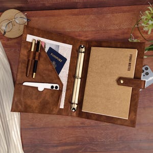 May include: A brown faux leather binder featuring a gold-toned three-ring mechanism, a document pocket containing a passport, two pen loops with pens, and a notebook labeled FENBI NOTES secured by a snap-button strap on the right side.