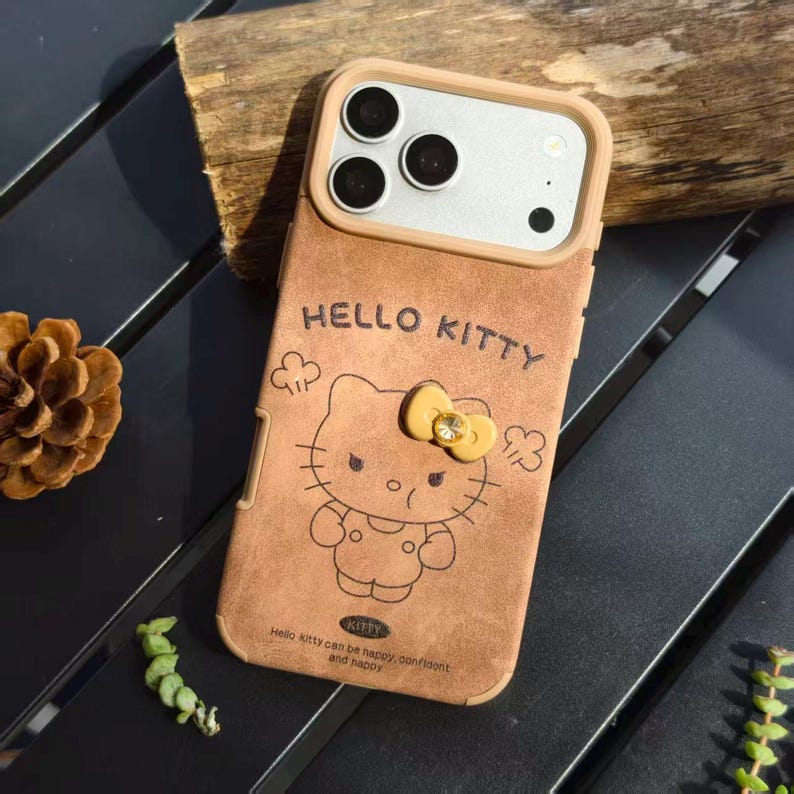 May include: Brown phone case featuring a Hello Kitty design. The case has a tan border around the camera and a Hello Kitty graphic with a yellow bow. The text "HELLO KITTY" is above the character, and the text "Hello Kitty can be happy, confident and happy" is below.