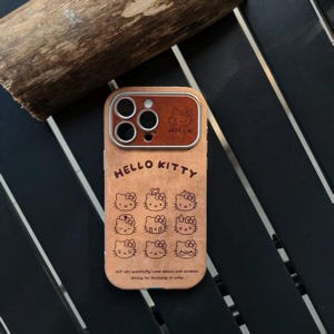 Op de afbeelding: Bruine telefoonhoes met Hello Kitty-ontwerp. De hoes heeft een bruine, leerachtige textuur met meerdere Hello Kitty gezichtsillustraties en de tekst "HELLO KITTY". De tekst "WO will eventoofly come ashore coth sonahios shining for thosands of wifes" is ook aanwezig.