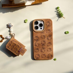 Peut inclure: Un &eacute;tui de t&eacute;l&eacute;phone marron avec un motif Hello Kitty et un porte-cartes assorti. L'&eacute;tui de t&eacute;l&eacute;phone a un bord beige autour des lentilles de la cam&eacute;ra. Le porte-cartes porte l'inscription "Hello Kitty".