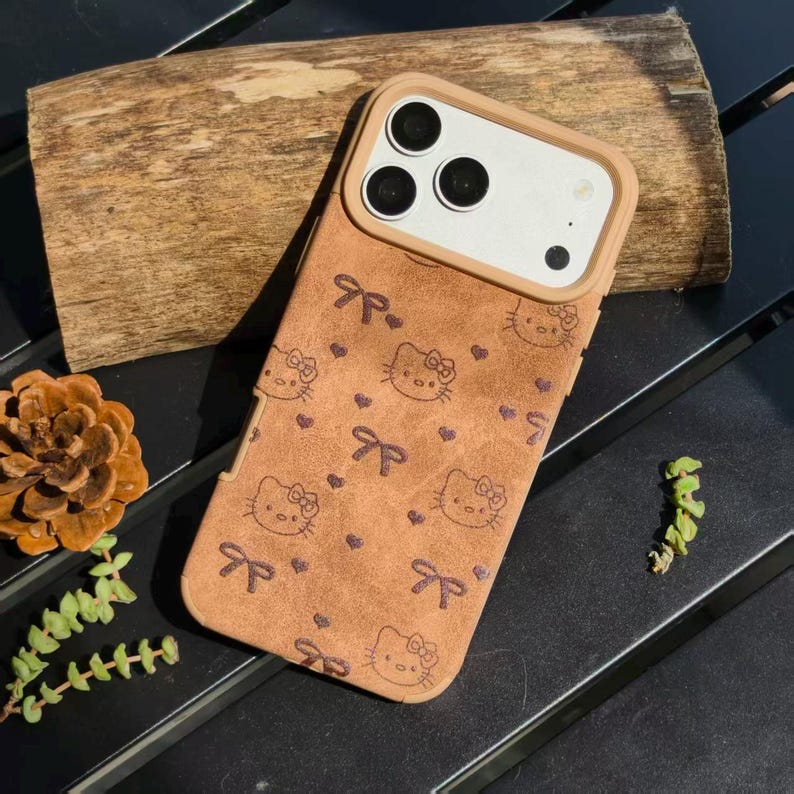 May include: A brown phone case featuring a repeating pattern of Hello Kitty faces, bows, and hearts. The case has a tan border around the camera and a matching tan top. The case is on a dark surface with a piece of wood.