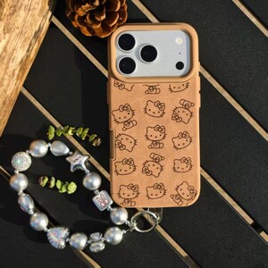 May include: Tan phone case with a repeating pattern of Hello Kitty characters. The case has a light brown border and a silver camera module. A beaded bracelet with silver and pearl-like beads and a pink star charm is next to the phone.