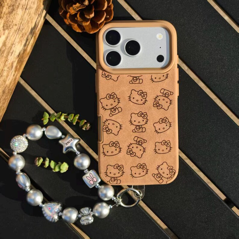 May include: Tan phone case with a repeating pattern of Hello Kitty characters. The case has a light brown border and a silver camera module. A beaded bracelet with silver and pearl-like beads and a pink star charm is next to the phone.