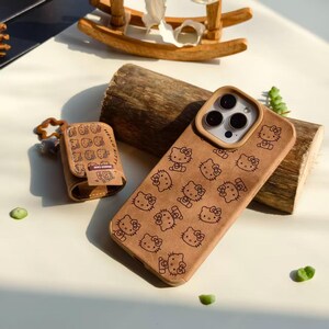 Peut inclure: Un &eacute;tui de t&eacute;l&eacute;phone marron et un &eacute;tui AirPod assorti, tous deux orn&eacute;s d'un motif r&eacute;p&eacute;t&eacute; de personnages Hello Kitty. Les &eacute;tuis sont fabriqu&eacute;s dans un mat&eacute;riau textur&eacute; et sont pos&eacute;s sur une surface claire, avec une b&ucirc;che de bois et un cheval &agrave; bascule jouet en arri&egrave;re-plan.