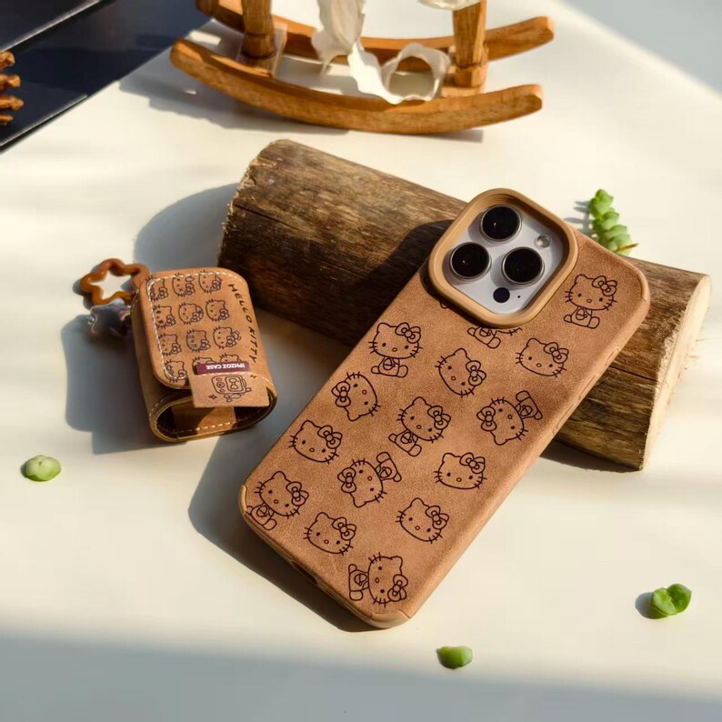 Peut inclure: Un &eacute;tui de t&eacute;l&eacute;phone marron et un &eacute;tui AirPod assorti, tous deux orn&eacute;s d'un motif r&eacute;p&eacute;t&eacute; de personnages Hello Kitty. Les &eacute;tuis sont fabriqu&eacute;s dans un mat&eacute;riau textur&eacute; et sont pos&eacute;s sur une surface claire, avec une b&ucirc;che de bois et un cheval &agrave; bascule jouet en arri&egrave;re-plan.
