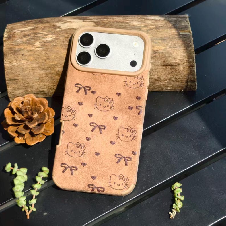 May include: Tan phone case featuring a repeating pattern of Hello Kitty faces, bows, and hearts. The case has a beige border and a silver camera module. The case is designed for a smartphone.