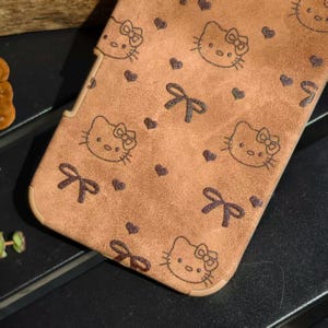 May include: A brown phone case featuring a repeating pattern of cartoon cat faces, bows, and hearts. The case has a textured appearance and a light brown border. The design is in a darker brown color.