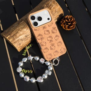May include: A tan phone case featuring a repeating pattern of Hello Kitty characters. A silver and pearl bracelet with charms and a silver clasp is next to the phone case. A piece of wood and a pinecone are also in the image.