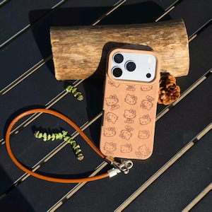 May include: A tan phone case featuring a repeating laser-engraved pattern of the Hello Kitty character. The case includes a matching tan leather-like wrist strap attached with silver-toned metal hardware and a raised camera lens protector frame.