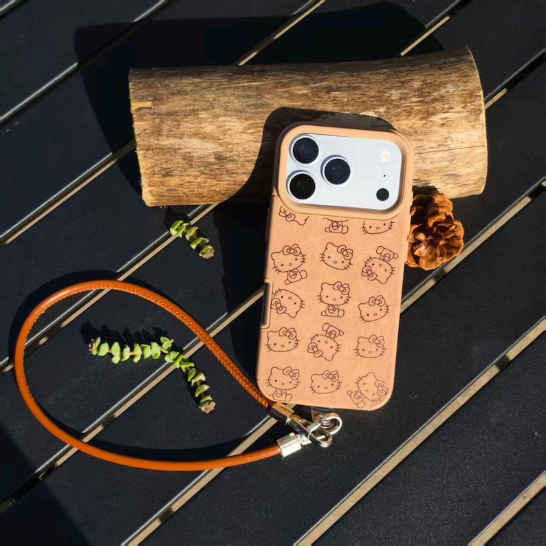 May include: A tan phone case featuring a repeating laser-engraved pattern of the Hello Kitty character. The case includes a matching tan leather-like wrist strap attached with silver-toned metal hardware and a raised camera lens protector frame.