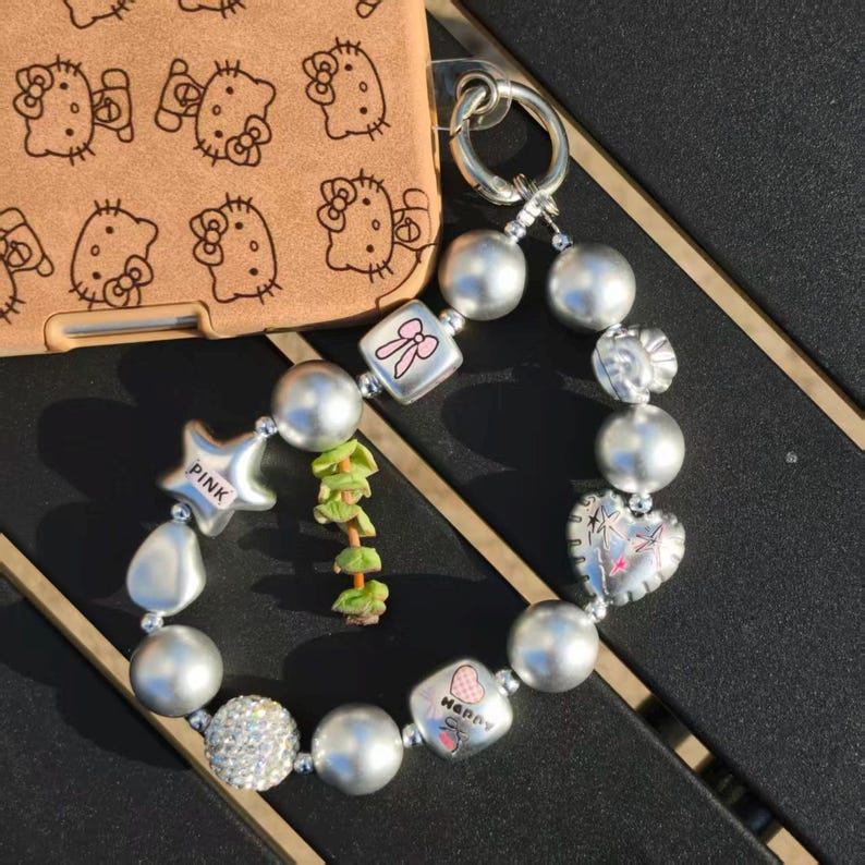 May include: Silver beaded bracelet with charms, including a star, heart, and bow. The bracelet features a mix of round and irregular beads, with a sparkling rhinestone accent bead. A brown phone case with a Hello Kitty pattern is in the background.