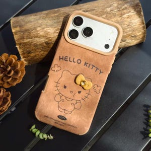 May include: Brown phone case featuring a Hello Kitty design. The case has a tan border around the camera and a Hello Kitty graphic with a yellow bow. The text "HELLO KITTY" is above the character, and the text "Hello Kitty can be happy, confident, and happy" is below.