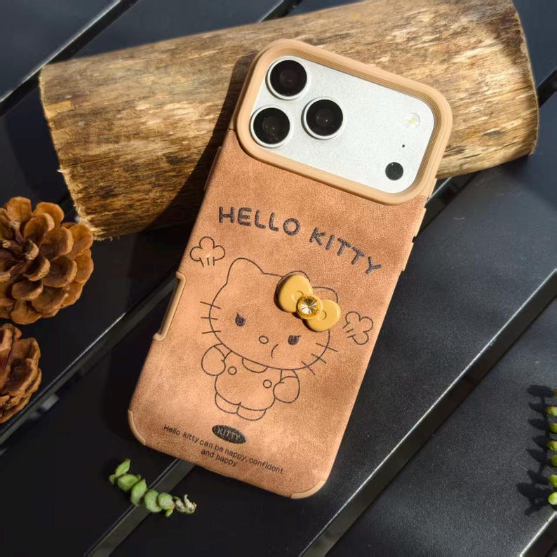 May include: Brown phone case featuring a Hello Kitty design. The case has a tan border around the camera and a Hello Kitty graphic with a yellow bow. The text "HELLO KITTY" is above the character, and the text "Hello Kitty can be happy, confident, and happy" is below.