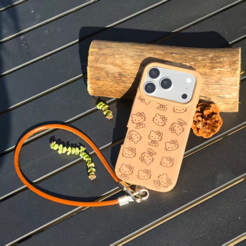 May include: A tan smartphone case featuring a repeating black line art pattern of the Hello Kitty character. The case includes a matching tan wrist strap attached with a silver metal clasp and a silver camera lens frame cutout.