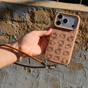 May include: A tan smartphone case featuring a repeating black line art pattern of the Hello Kitty character. The case includes a raised camera frame and a thin, brown leather-like wrist strap attached with a silver-toned metal clasp.
